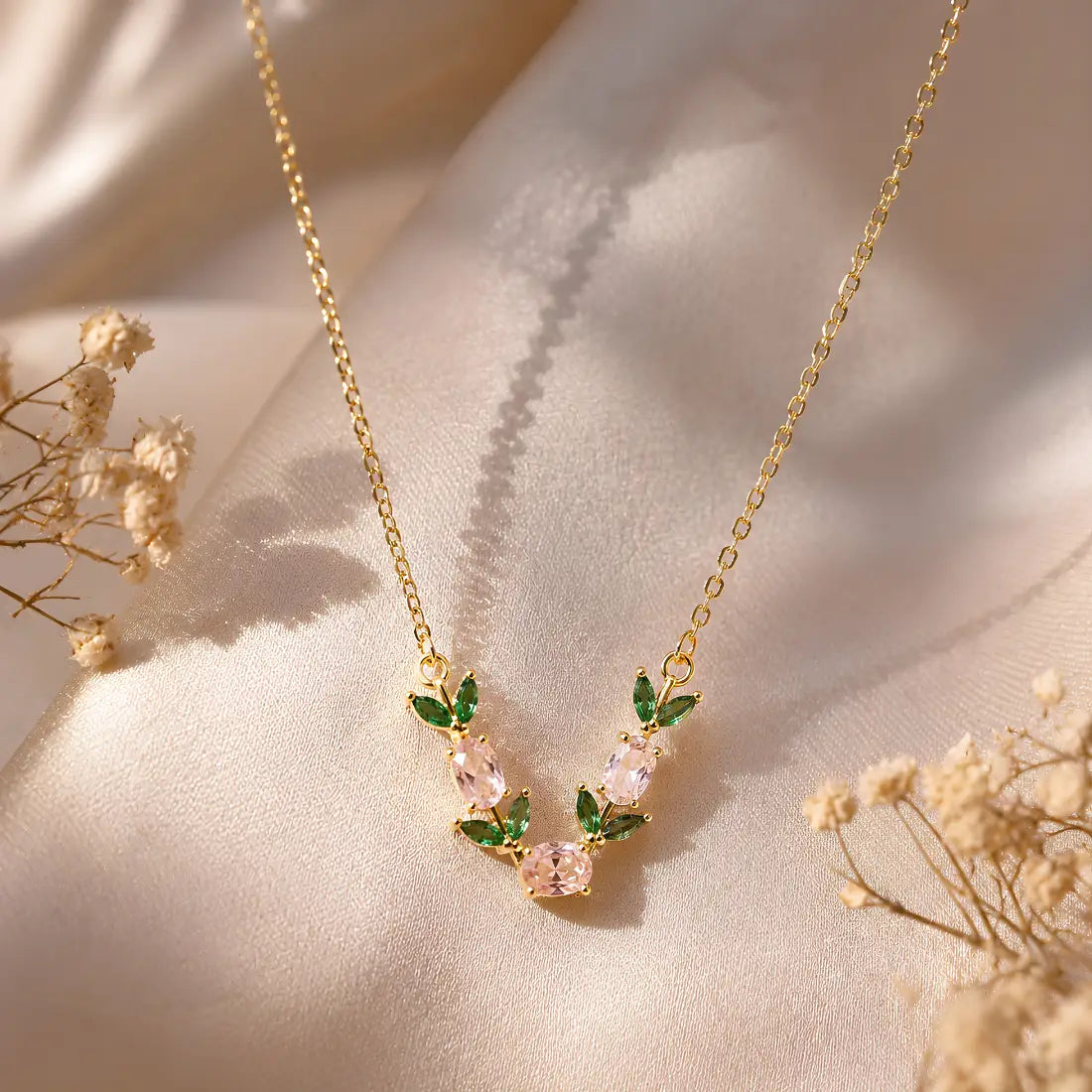 Gold necklace with three pink oval gemstones and green marquise gemstones arranged in a leafy vine design, shown on a light cream background.